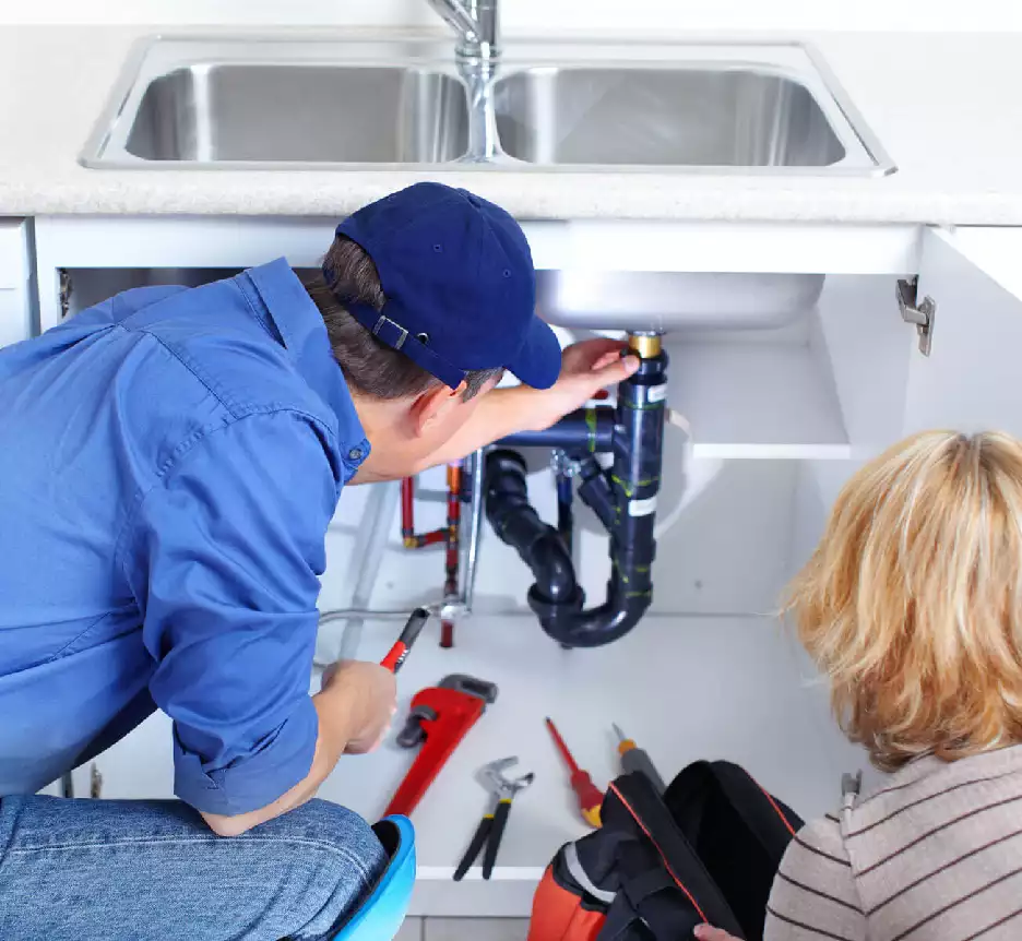 Picture of a plumber working on the plumbing under a sink with the homeowner observing.